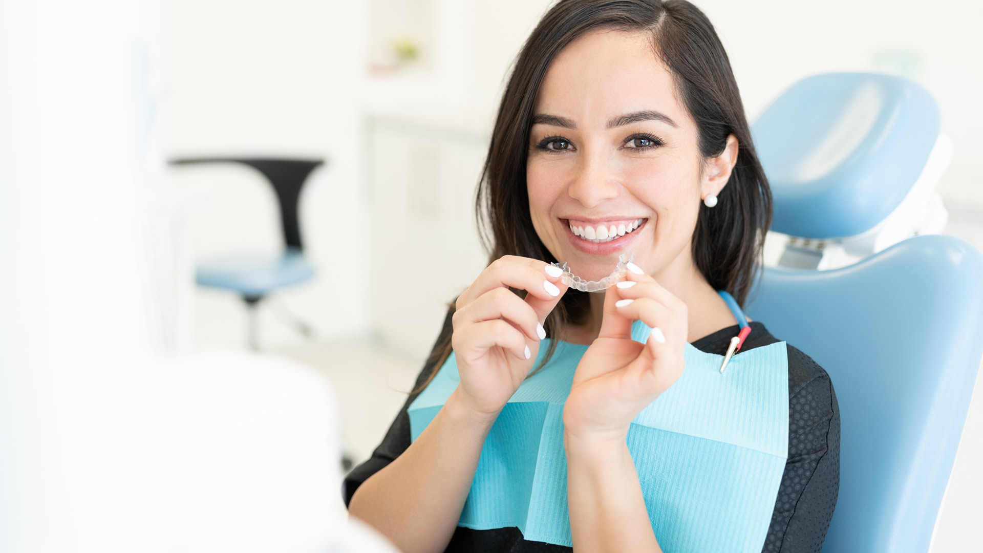 Female patient smiling in dental chair while holding Invisalign clear aligner at SS Orthodontics in Glendale and Phoenix, AZ